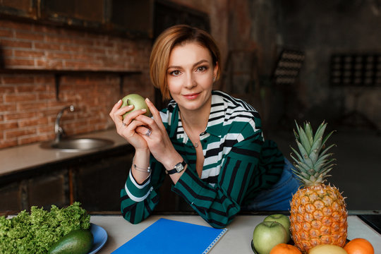 Beautiful young woman holding apple and looking to camera in her modern loft kitchen. Fitness coach. Healthy food concept.