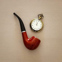 Top view of old smoking pipe and vintage pocket watch over wooden background.