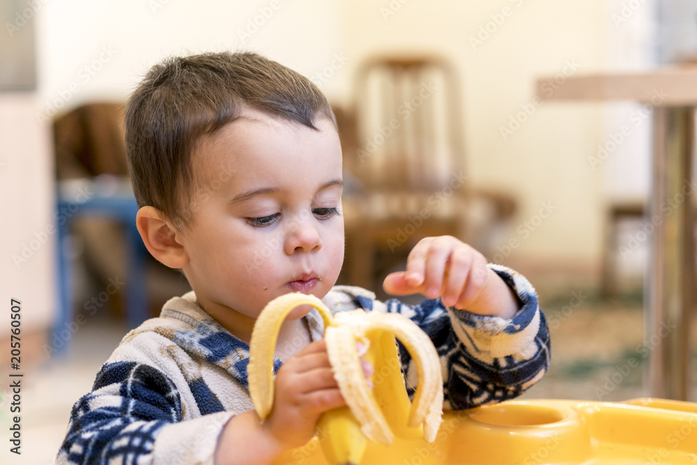 Wall mural Funny boy 2 years old eating banana. Funny boy 2 years old eating banana