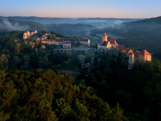 Aerial view of beautiful, Moravian royal castle Veveri or Burg Eichhorn, standing on a rock above water dam on river Svratka. Large castle above misty trees in early morning light. Aerial photography. - obrazy, fototapety, plakaty