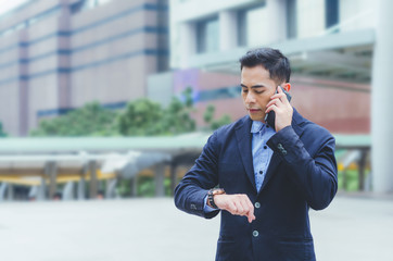 portrait attractive handsome business man using smartphone talking and looking smart watch at outdoor city with building background. technology connecting social online .