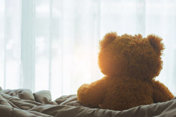 brown teddy bear sitting on bed looking on windows with sunlight in the bedroom on morning.