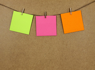 Hojas de papel de color verde, naranja  y rosa (posit), sujeto con una pequeña pinza de madera y una cuerda a una pared de corcho. 