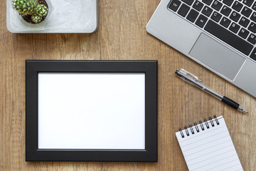 A wooden desk with a laptop, a cactus in a concrete pot, a notebook and a black empty frame