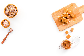Cook caramel sauce. Melted caramel in glass jar, caramel cubes on cutting board, sugar and salt on white background top view space for text