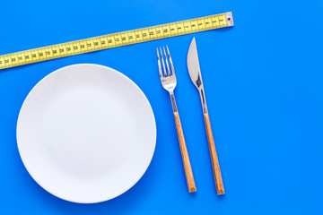 Diet for weight loss concept. Proper nutrition. Medical starvation. Empty plate with fork and knife near measuring tape on blue background top view mockup
