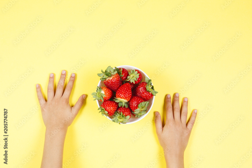 Wall mural kid hands and bowl with fresh strawberries on yellow background.