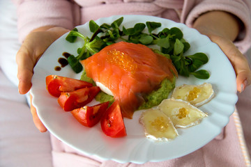 White plate with healthy meal include smoked salmon, avocado; vegan seaweed pearls caviar; goat cheese; fresh tomato and lettuce, woman hands holding the plate with meal in bed
