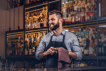 Cheerful stylish brutal barman is cleaning the glass with a cloth at bar counter background. - Powered by Adobe