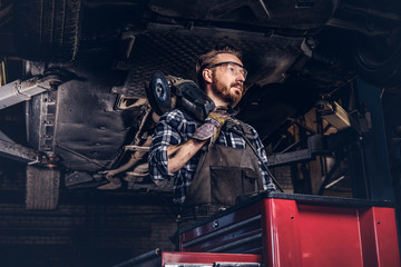 Fototapeta na wymiar Mechanic in a uniform and safety glasses holds an angle grinder while standing under lifting car in a repair garage. 