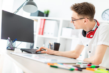A young man in glasses stands near a computer desk. A young man works with a computer and a magnetic board. On the neck, the guy's headphones hang.