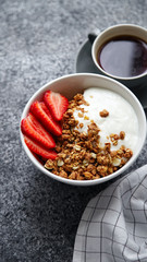 Close up shot of perfect breakfast: crunchy granola with yoghurt and strawberries with a cup of black coffee on gray bakcground.