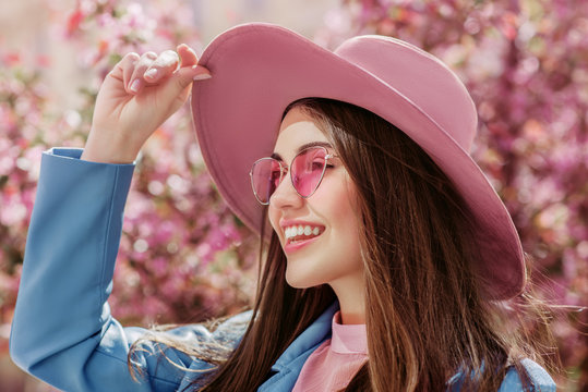 Outdoor close up portrait of young beautiful happy smiling girl wearing trendy pink color sunglasses, suede  hat, blue jacket. Model looking aside. Spring, summer fashion concept
