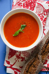 Tomato soup garnished with basil leaves served with rye bread on blue wooden table