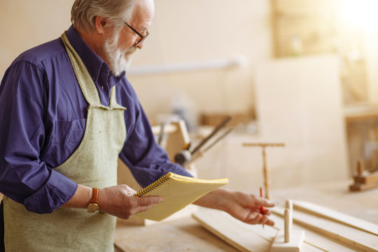 old man in shirt and apron is measuring the plank in the capenter's workshop. closeup side view shot