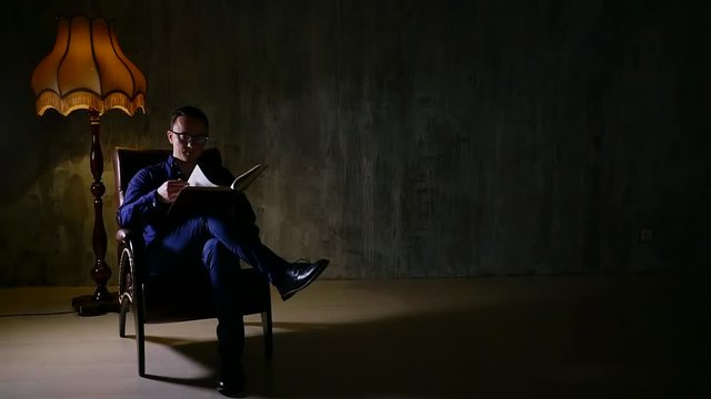 Serious man is sitting in a chair wearing suit and reading a book under the lamp.