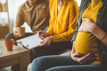 Focus on pregnant woman touching her abdomen with gentleness. Married couple is putting signature on document while sitting on background. Surrogate motherhood concept 