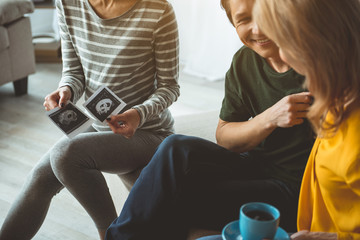 Happy future parents are observing fetus of pregnant woman on photo. They are sitting near the surrogate expectant mother and smiling 