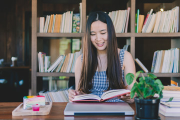 Beautiful Young Asian Woman sitting and reading book in library with book shelf in background, cake, laptop and flower on wood table