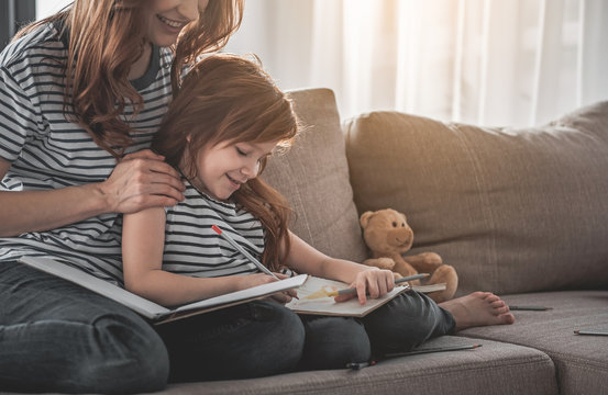 Family harmony. Portrait of smiling kid sitting on sofa with her mom holding her shoulders. Red-haired woman is watching her daughter with content. Pencils are scattered on sofa beside them