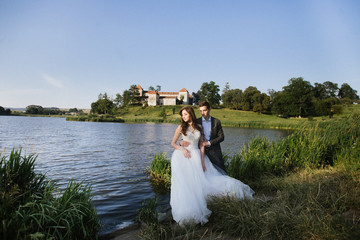 happy wedding couple walking holding hands in evening field on background of old castle. elegant bride and groom embracing. romantic moment. man in suit with bow tie and woman in dress with pearls
