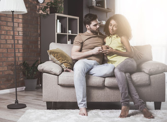 Obraz premium Strong relationship. Portrait of charming girl is resting on cozy sofa with her boyfriend. She is looking at camera with smile while man is watching her with adoration. They are holding cup together