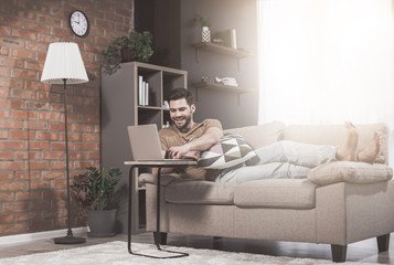 Freelancer. Full length of optimistic young bristled man is lying on cozy sofa while working at home. He is using modern laptop with smile. Sunlight in background