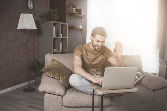 Hi there. Full-length portrait of cheerful guy is sitting on cozy sofa and enjoying video call on his modern laptop. He is emotionally waving his hand while talking in net. Sunlight in background