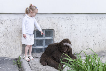 Little beautiful girl standing at the wall of the house points to a large toy monkey sitting next - Powered by Adobe
