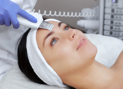 The doctor-cosmetologist makes the apparatus a procedure of ultrasound cleaning of the facial skin of a beautiful, young woman in a beauty salon. Cosmetology and professional skin care.