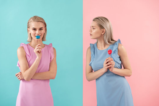 Two girls holding sweets on stick. One looking at another with envy. Isolated on blue and pink background