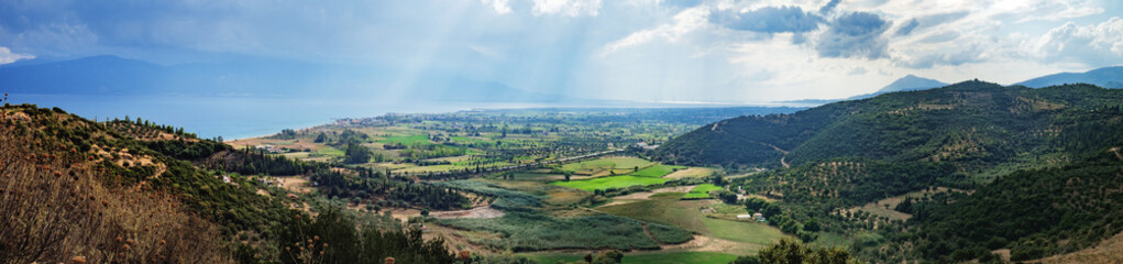 Summer rural landscape of Western Greece. Green forest on hillsides and house with red roofs on shore of the Gulf of Corinth. Rion-Antirion Bridge is seen in the distance