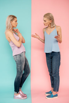Sibling rivalry. Full length of two discontented sisters talking and gesticulating while standing on blue and pink background