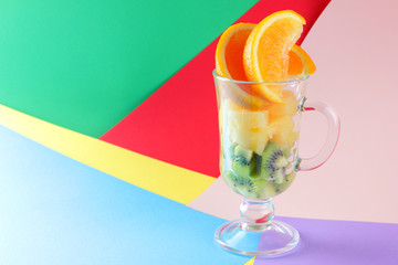 Pieces of tropical fruits in a glass, fruit cocktail. Sliced orange, kiwi cubes, slices of pineapple in a glass bowl. Multi-colored pop art background, copy space