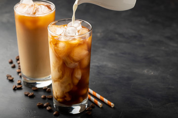 Ice coffee in a tall glass with cream poured over, brown sugar and coffee beans. Cold summer drink on a dark background. With copy space