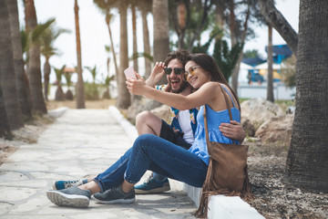 Joyful young guy and girl are making selfie in tropical park. They are sitting on the path and hugging while laughing 