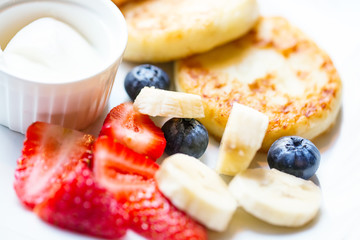 Cheesecakes syrniki on white plate with banana pieces, strawberry and blueberry on a plate with sour cream. Close up image with selective focus.