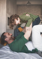 Side view cheerful dad keeping laughing kid in hands while lying on bed