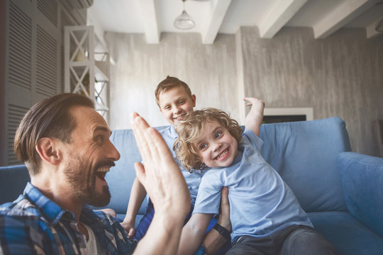 Outgoing Child Giving High Five For Laughing Dad. Cheerful Elder Brother Sitting Near Him. Satisfied Father And Glad Children Gesticulating Hands Concept