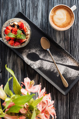 top view of tasty healthy dessert with fruits, spoon, cup of coffee and flowers on wooden table