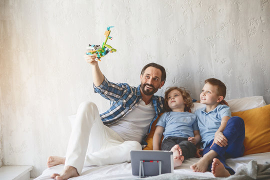 Full length portrait of cheerful kids having good time with pleased dad while sitting on bed
