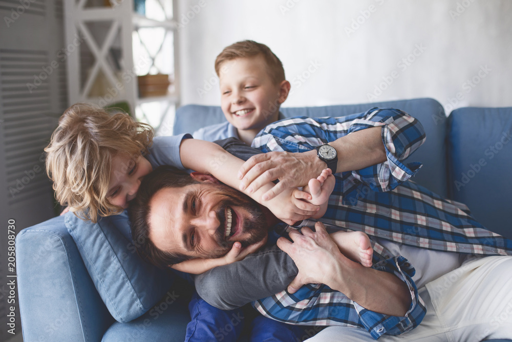 Wall mural Portrait of smiling father and outgoing sons having fun on cozy couch in apartment. Happy family concept