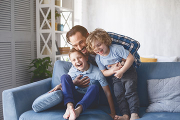 Portrait of laughing dad hugging beaming kids having entertainment on sofa in living room....