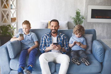 Obraz premium Portrait of cheerful bearded dad sitting between two smiling children on cozy sofa. They using gadgets. Happy family concept