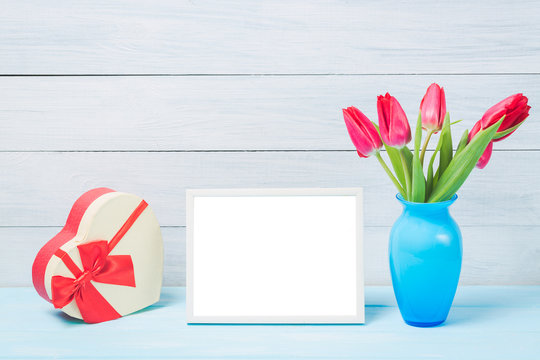 Colorful red spring tulip flowers in nice blue vase and blank photo frame with decorative heart giftbox on light wooden background as greeting card. Mothersday or spring concept