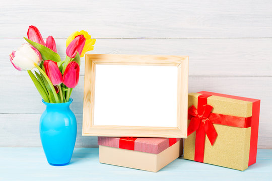 Colorful red spring tulip flowers in nice blue vase and blank photo frame with giftboxes on light wooden background as greeting card. Mothersday or spring concept