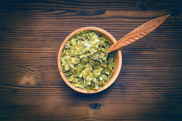 Green dry hop leaves in wooden bowl and spoon isolated on brown table background.