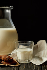 partial view of fresh milk in jug and glass, chocolate cookies and sackcloth on black background
