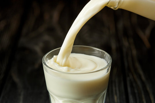 Milk Pouring Into Glass From Bottle On Black Wooden Surface