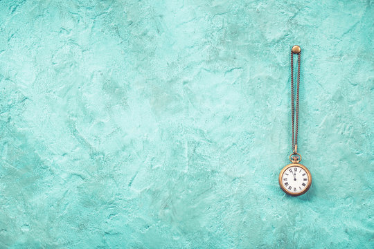 Vintage bronze pocket watches with five minutes to twelve o'clock hanging on nail front old mint green concrete wall textured background. Retro style filtered photo